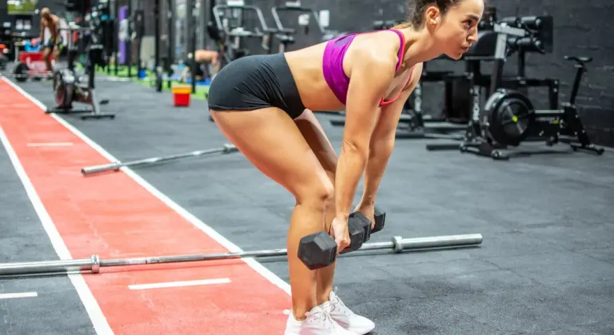 Mujer concentrada realizando un **double db deadlift** en un gimnasio, con pesas en cada mano y el cuerpo inclinado.