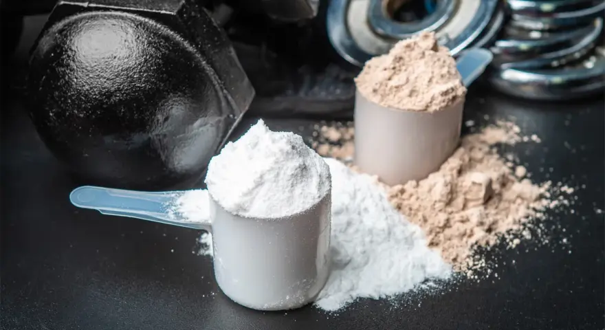 Dos cucharadas de proteína en polvo, una blanca y otra marrón, junto a pesas. ¡Cuidado! la proteína en polvo caduca.