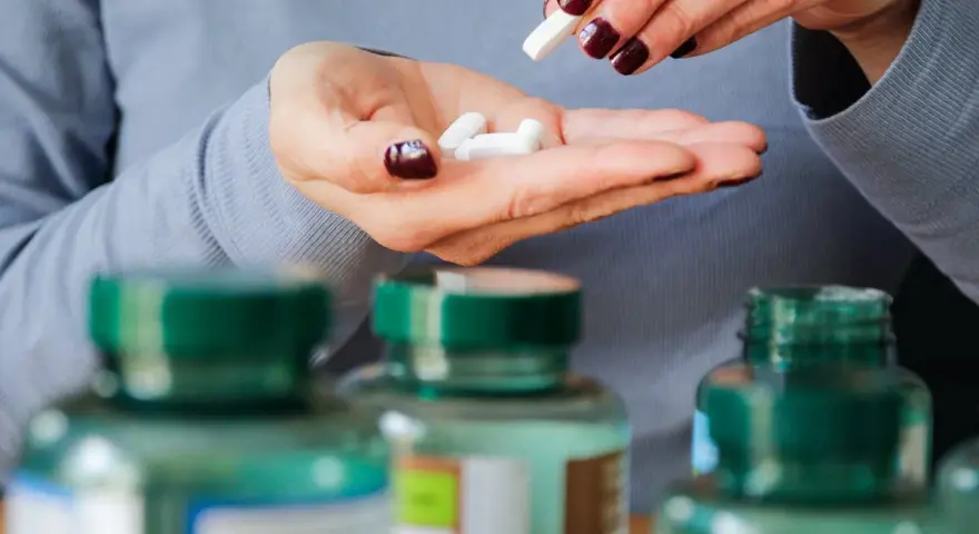 Manos con uñas oscuras sostienen pastillas blancas. Se ven frascos de suplementos. Descubre los beneficios del magnesio en el hombre.