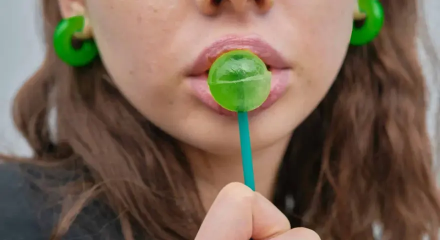 Labios rosados con un chupachups verde. Los pendientes son grandes y verdes. El maltitol es malo, pero este dulce parece tentador.
