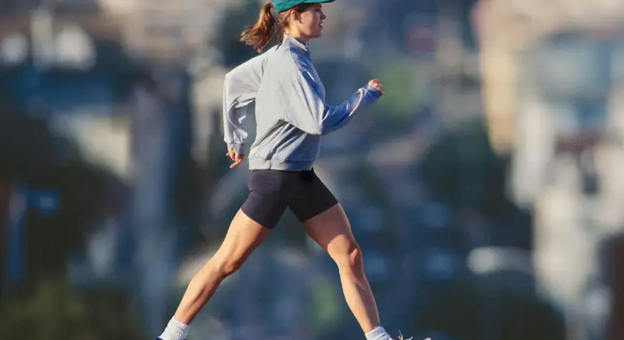 Mujer con gorra y sudadera gris corriendo al aire libre, disfrutando de un paseo después de comer para mantenerse activa.