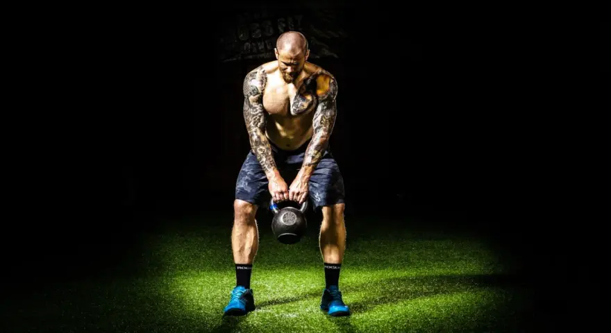 Hombre tatuado realiza press militar con kettlebell, enfocado en su entrenamiento.