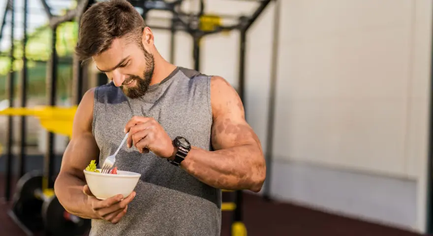 Hombre con barba en gimnasio, comiendo ensalada. Demuestra que se puede ganar músculo en déficit calórico con la dieta adecuada.