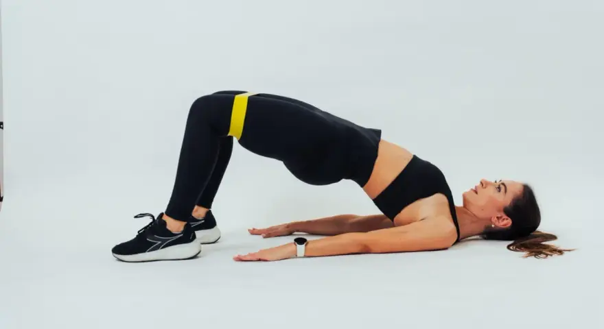 Mujer con banda amarilla en glúteos, en posición de glute bridge hold, con fondo blanco.