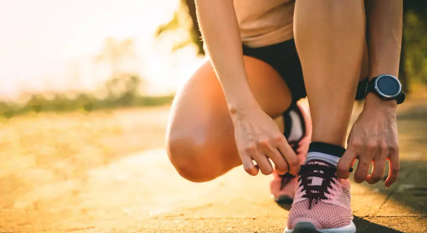 Persona se agacha para atarse los cordones de sus zapatillas rosas, lista para salir a correr al amanecer.