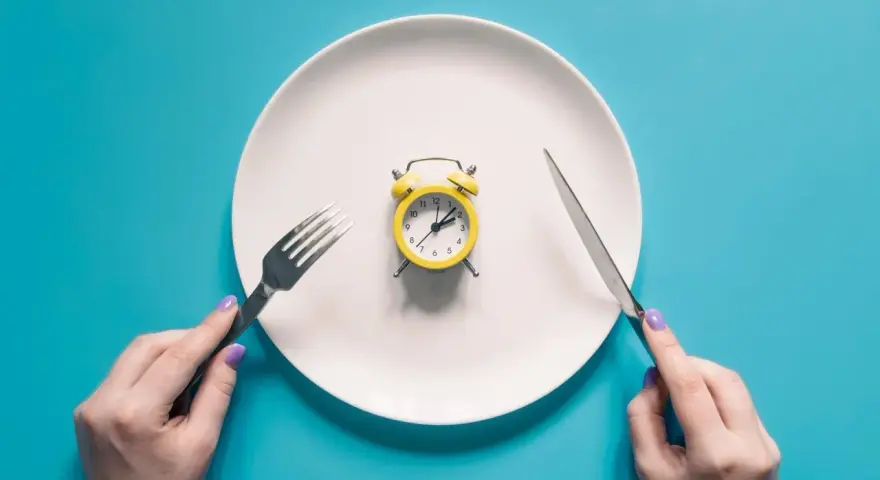 Plato blanco con despertador amarillo en el centro. Manos sostienen tenedor y cuchillo. El café rompe el ayuno.