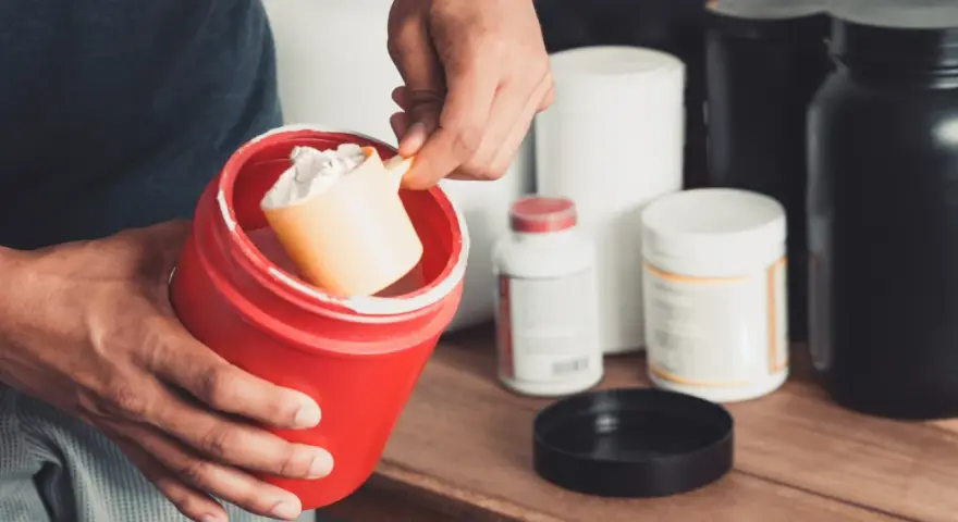Persona prepara su suplemento. Se muestra un recipiente rojo con polvo blanco y una cuchara. Varios envases de suplementos en el fondo. Calculando la dosis creatina por peso.