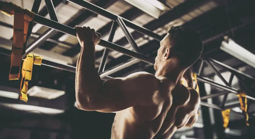 Hombre musculoso realizando pull ups en un gimnasio. Sus brazos y espalda están tensos mientras se eleva.