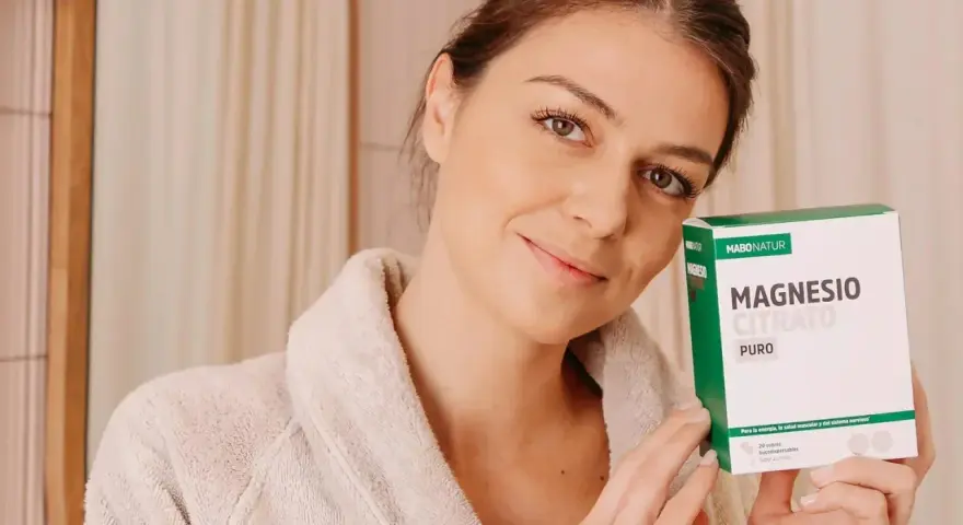 Mujer sonriente con bata, sosteniendo una caja de Magnesio Citrato Puro. Una dosis de magnesio para evitar calambres y mejorar la energía.