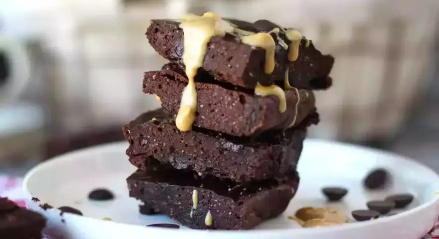 Apilados trozos de brownie de proteína cubiertos con salsa de caramelo y chispas de chocolate.