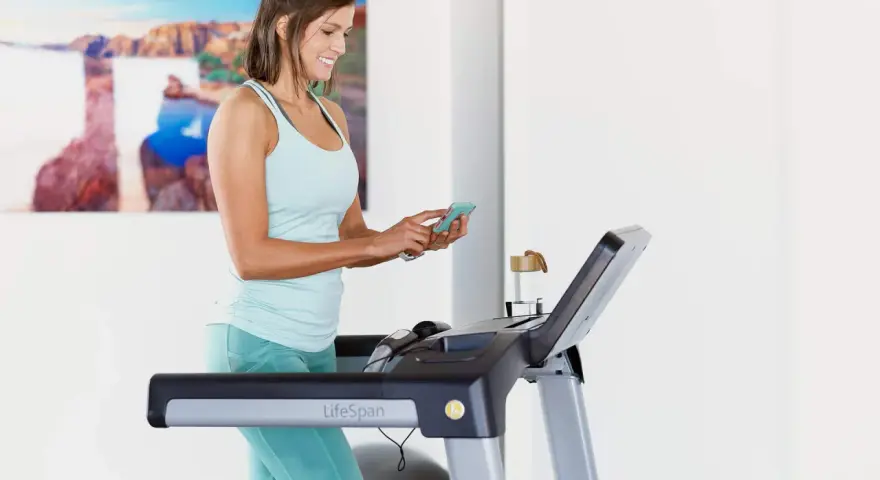 Mujer sonriente en cinta de correr, revisando su teléfono. Quizás buscando cuántas calorías debo comer al día para mantener su energía.