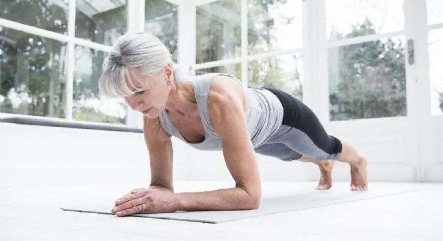 Mujer mayor haciendo plancha en su sala, demostrando que el fitness en casa es para todas las edades.