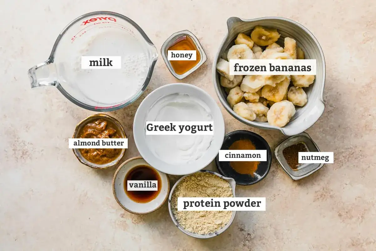 Ingredientes para un batido de proteínas casero: leche, yogur griego, plátanos congelados, mantequilla de almendras, miel, vainilla, canela y nuez moscada.