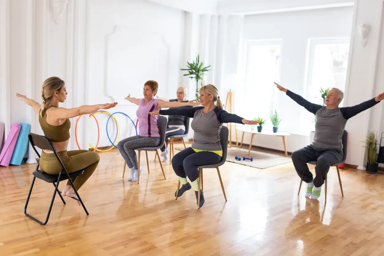 Grupo de personas mayores realizando ejercicios en silla, guiados por una instructora.