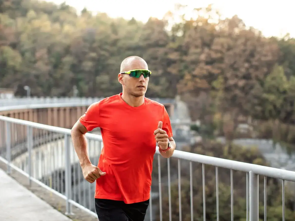 Hombre corriendo con gafas de correr deportivas, camiseta naranja y fondo de naturaleza.