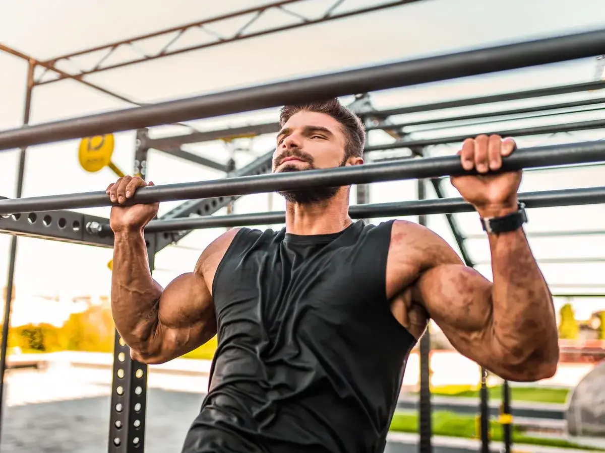 Hombre musculoso haciendo pull ups en una barra exterior. Sus brazos est&aacute;n tensos y sus m&uacute;sculos definidos.