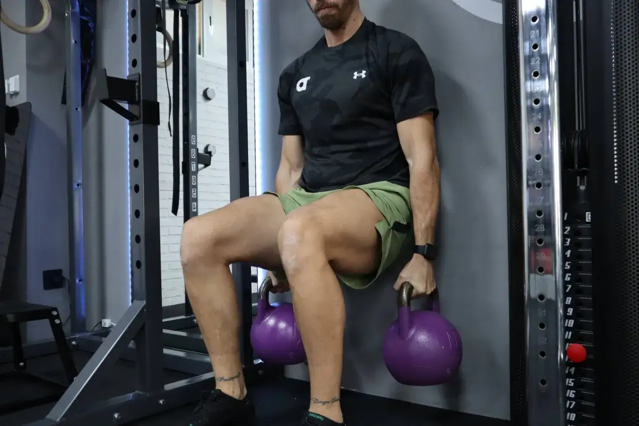 Hombre realizando sentadilla isométrica en pared con pesas rusas. Una tabla de ejercicios isométricos es ideal para este entrenamiento.