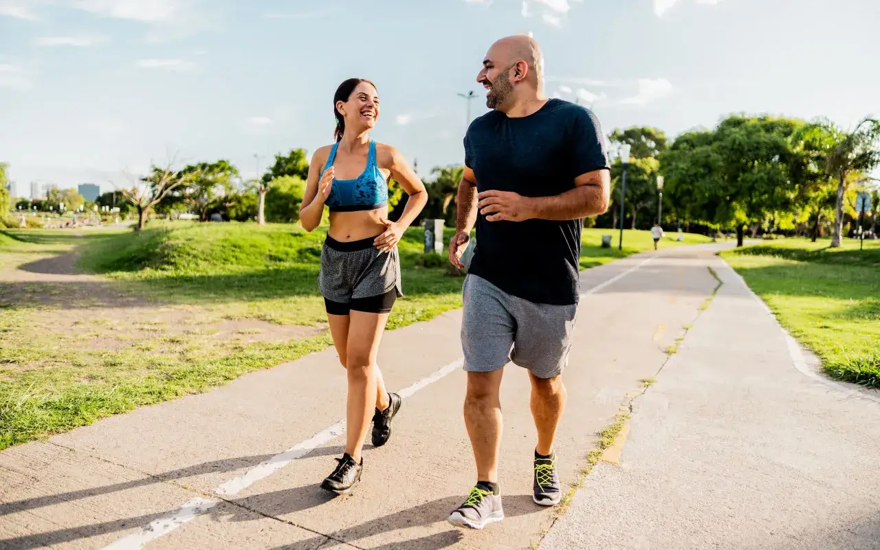 Pareja sonríe mientras corre por un parque. El andar adelgaza y les ayuda a mantenerse en forma.