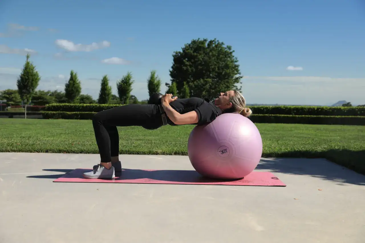 Mujer haciendo puente con pesa rusa sobre pelota de ejercicio, un ejercicio ideal para como activar las mitocondrias y mejorar la energ&iacute;a.
