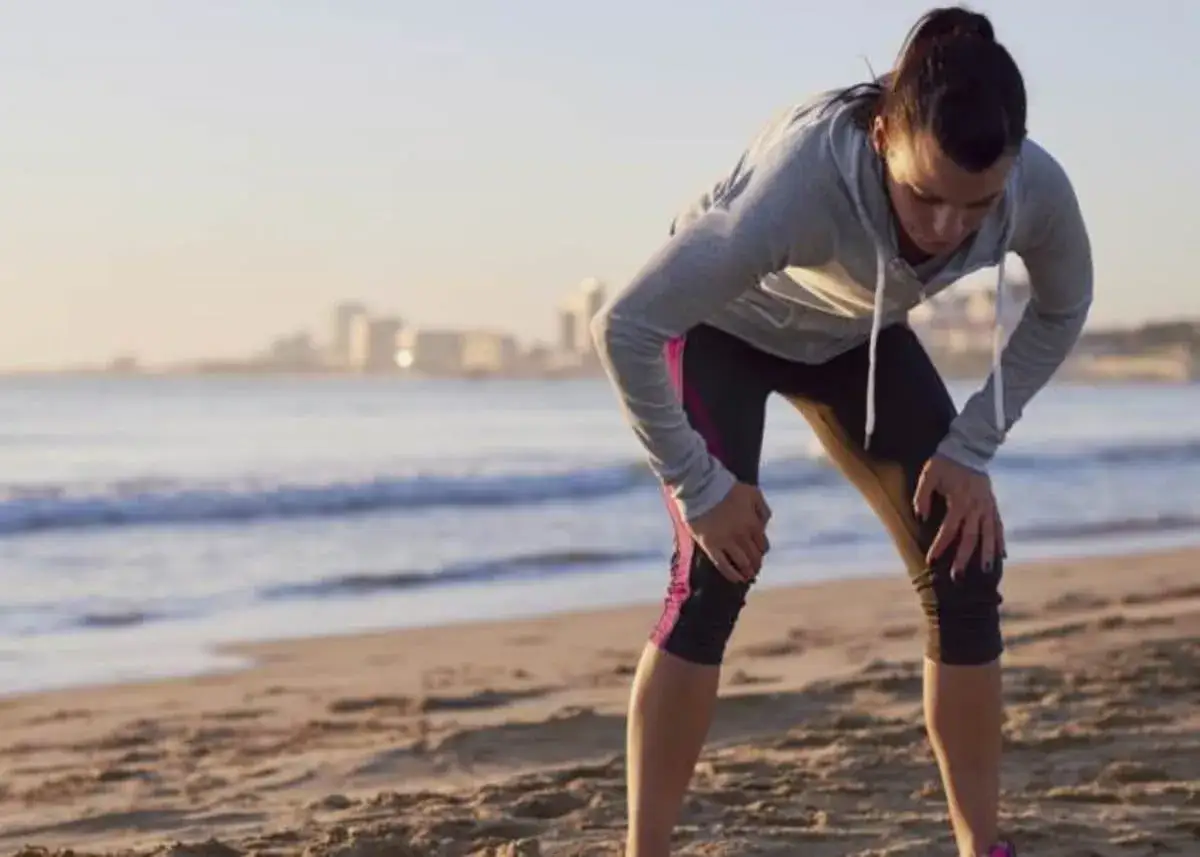 Corredora en la playa, con las manos en las rodillas, mostrando síntomas de cansancio y sueño tras un entrenamiento.
