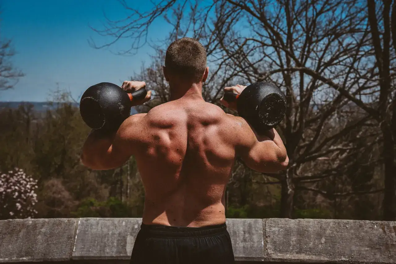 Hombre musculoso realiza press militar con kettlebell, entrenando al aire libre con &aacute;rboles de fondo.